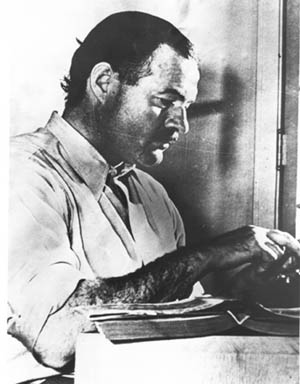Dark-haired man in light colored short-sleeved shirt working on a typewriter at a table on which sits an open book