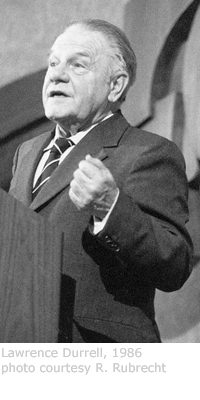 Durrell stands proud at a podium, gazing at the crowd as he addresses them. He wears a suit over a white shirt with a striped tie, and holds his left arm at his side, with his elbow bent upwards as if to shake his fist. A caption runs below the image that reads "Lawrence Durrell, 1986 - photo courtesy R. Rubrecht."