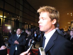 A Caucasian male with dyed blonde hair is being interviewed. He is wearing a black suit and tie, with a white shirt, and is standing on a red carpet. People standing behind barricades are visible in the background, while microphones are visible in the foreground.