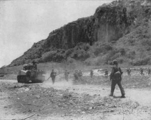 tank in mid-left heading down road from left to right. Terrain is flat in foreground, forested cliffs in back ground. Soldiers are walking behind the tank