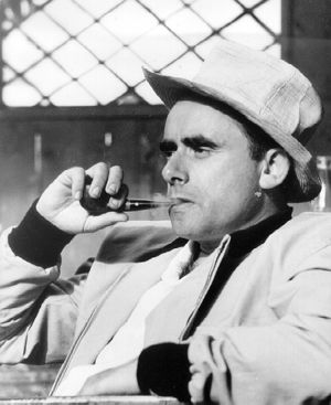 A black-and-white photo of Henri-Georges Clouzot taken from a side. Clouzot is depicted sitting downsmoking a pipe and wearing a matching white and hat and suit.