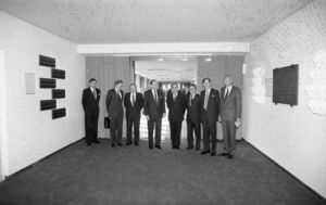 Eight men in suits stand in a hall facing forward.