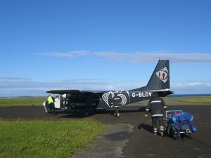 A small dark-blue twin prop plane sits on tarmac surrounded by grass under blue skies. In the foreground an individual wearing a uniform that is similar in colour to the plane pulls a full baggage trolley towards it.