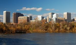 Numerous high-rise buildings rises over a dark river and its leafy autumnal shore.