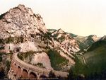 A photochorm picture of the a curving railroad bridge coming out of a tunnel bored into a large mountain. In the background another bridge and mountains are visible.