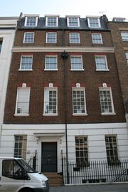 A terrace house with four floors and an attic. It is red brick, with a slate roof, and the ground floor rendered in imitation of stone and painted white. Each upper floor has four sash windows, divided into small panes. The door, with a canopy over it, occupies the place of the second window from the left on the ground floor.