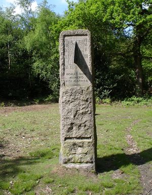 A standing stone in a grassy field surrounded by trees. The stone contains a vertical sundial centered on 1 o'clock, and is inscribed "HORAS NON NUMERO NISI ÆSTIVAS" and "SUMMER TIME ACT 1925".