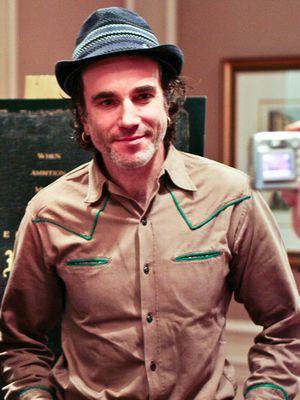 A smiling man wearing a grey hat with piping above the band, and a tan Western style shirt, stands in an office, posing for the camera.