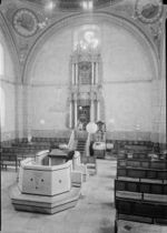 A sepia photograph shows the interior of the synagogue. In the foreground lies the reading desk atop a simply paneled almemar. Rows of wooden benches line the right and left side of the nave. The holy ark, set in the centre of the eastern wall, is decorated with baroque carvings and set off against four Corinthian columns. Its top reaches a large clover shaped window which sits just below one of the four supporting arches. The walls faintly show decorative murals, with two large round frescos situated at the top left and right corners.
