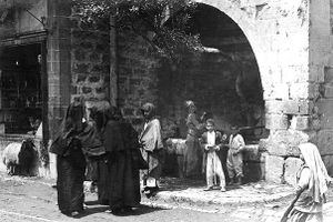 From left to right: A boy staring out from a store's window sill beneath which a cobra is walking by; three fully clothed women are conversing on the street; beneath an olive grove jutting out of a large stone archway and beside a fountain, a man is walking, a woman is collecting water from the fountain, and two young boys are standing and smiling; a young girl walking on the street