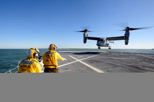 A V-22 performing a vertical landing on USS New York with two of the ship's crew nearby.