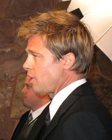 A side view of a Caucasian male, who is facing to the left, with light brown hair. He is wearing a black suit and tie with a white shirt. Another Caucasian male, also wearing a suit, is visible in the background.