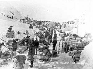 Peak of Chilkoot Pass in March–April 1898. Men wearing winter clothes with their supplies in the snow all of it surrounded by hill-sides.