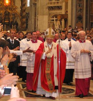 Pope Benedict XVI, wearing robes of red and white, is walking in procession in St. Peter's, and raising his hand in blessing.