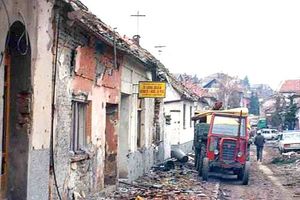 A street of ruined buildings with rubble strewn across the road. A red tractor and other vehicles are visible parked in the background