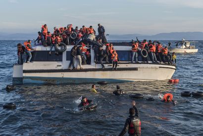 A small boat in water, with land on the horizon behind. Many people are on its outside in orange life jackets, some carrying inner tubes as well. A few are in the water swimming toward the camera. In the foreground a man in a red and back wetsuit has his hand out to them.