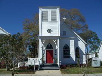 Small white clapboard church. The entrance is on the left, under another story just above the entrance, which gives the church a lopsided appearance.