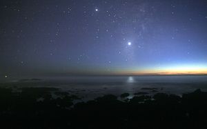 A photograph of the night sky taken from the seashore. A glimmer of sunlight is on the horizon. There are many stars visible. Venus is at the center, much brighter than any of the stars, and its light can be seen reflected in the ocean.