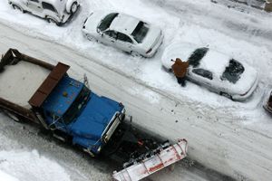 Street seen from above covered in snow with a city truck full of snow and a person who has to remove snow from his or her car