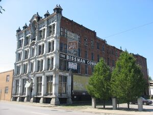 A large, red-brick building with a peeling, white-painted front: A sign on the side of the building is obscured by trees, but partially reads "The Bissman Comp..."
