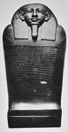 Black and white image of a dark stone coffin with a human face, the coffin stands upright facing the viewer.