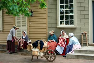 Five women dressed in long colonial style clothing sit on the stairs of tan and beige buildings talking. In front of them is a wooden wheelbarrow full of wicker baskets.