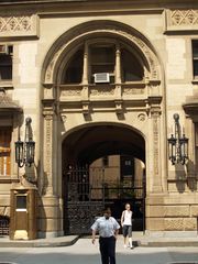 Exterior of a tan-coloured building, with a black metal gate in the middle of a prominent arch. A blue-clad policeman blowing a whistle and a woman are visible in front of the gate.