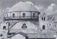 A large domed roof sits upon a bricked parapet, which includes arched windows, three of which are visible. The base of the parapet is surrounded by a narrow veranda which is fenced with decorative metal grating. Below this, a central clover shaped window lies at the top of an arched wall. Two supporting towers stand on the left and right, each containing an arched window. The right tower, capped with a small dome, is a storey higher, reaching to the top of the bricked parapet which supports the domed roof.