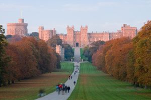 A red castle with battlements and towers lies in the distance of the photograph. A path curves from the bottom of the picture towards it, with various people strolling along it. On either side is flat grass and green woodlands.