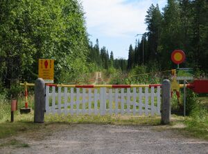 A simple white fence with a red and yellow gate behind it set across a dirt path in a green forest.