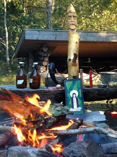 An outdoor fire burning in front of a wooden post with an anthropomorphic face carved into the top