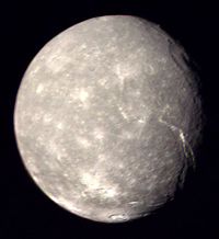 A round spherical body is almost fully illuminated by the Sun. The surface has a mottled appearance with bright patches among relatively dark terrain. The terminator is located near the right edge. A large crater can be seen at the terminator in the upper half of the image. Another bright crater can be seen at the bottom. A large canyon runs from the darkness at the lower-right side to visible center of the body.