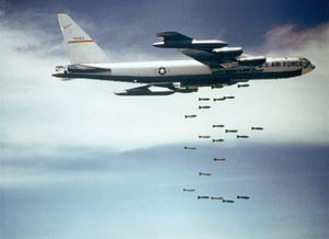 Against a blue sky with white clouds, a B-52F releases bombs over Vietnam.