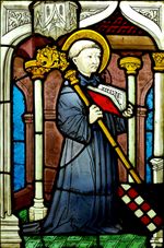 Stained glass image of a kneeling man with a halo holding an open book and a staff.