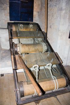 A rectangular metal frame with two wooden planks crossing its width. Spaced equally along the frame are three large wooden rollers. Attached to the rollers at each end are ropes, designed to act as restraints. The entire contraption is covered with a sheet of clear plastic, upon which is sketched the outline of a man, his wrists and ankles 'through' the rope restraints. The centre roller has a large wooden lever—turning this lever would pull the other two rollers in opposite directions.