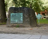 A rectangular, rough-hewn block of stone on the ground. There is a walkway in the foreground, grass and trees in the background. A weathered plaque on the stone reads: "Samuel Adams, Signer of the Declaration of Independence, Governor of this Commonwealth, A Leader of Men and an ardent Patriot."