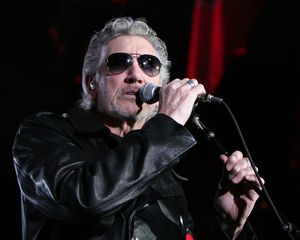 Waters on stage wearing sunglasses and a black leather coat. He is holding a microphone up to his mouth.
