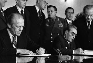 Two men in suits are seated, each signing a document in front of them. Six men, one in a military uniform, stand behind them.