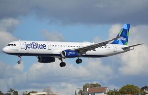 A white plane with the words "jetBlue" painted at the front and a blue-green tailfin approaches landing with its landing gear deployed as it soars above houses below