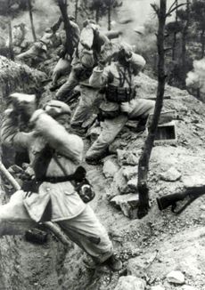 A line of soldiers are standing in front of a trench and holding rocks over their shoulders. Rocks are flying to the right while smokes are filling the background