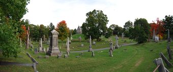 A panoramic view of the cemetery; a dirt road pass in front of the view from right to left. At center is a grey church-like structure, surrounded on all sides by various sized grave stones.