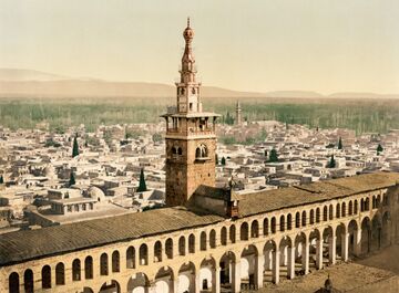 A color photochrom cityscape of 19th-century Damascus, showing a tower rising over an arcade in the forefront, old buildings in the background and gardens and hills on the horizon