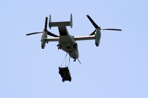 A MV-22 with its rotors up to vertical with a HMMWV vehicle hanging by two sling wires.